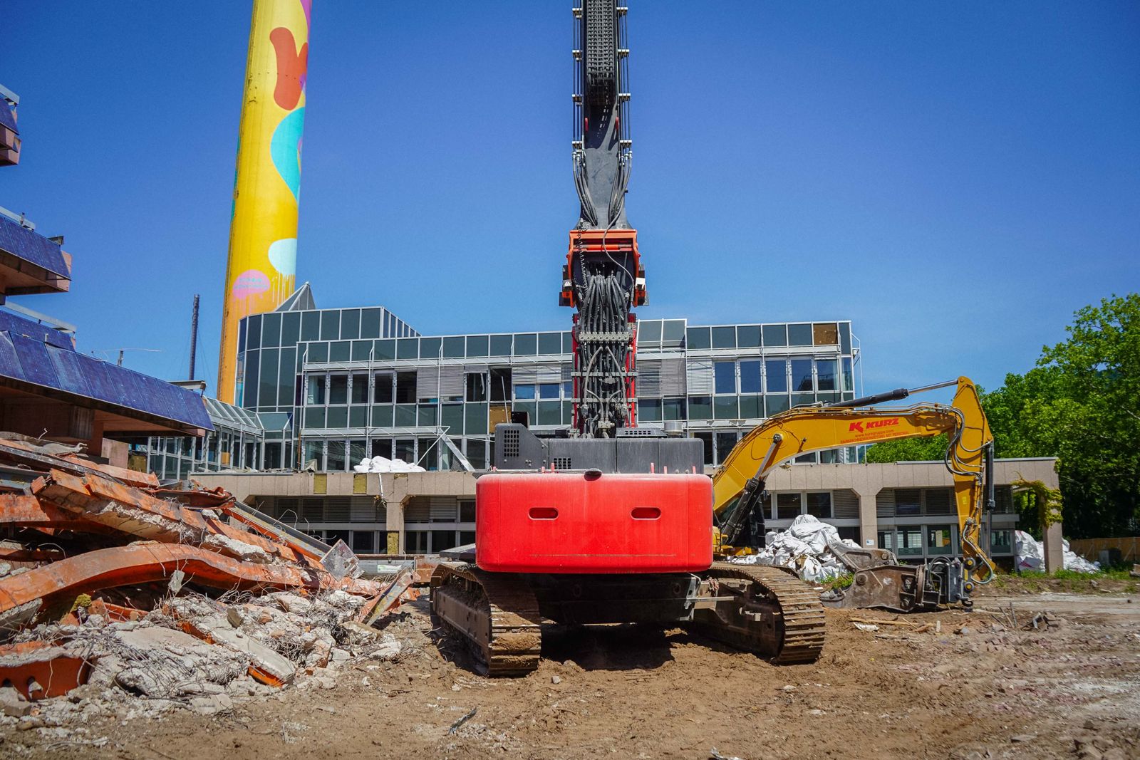 Roter Longfront-Abbruchbagger frontal auf einer Rueckbau-Baustelle mit Ziegelschutt und Glasgebaeude im Hintergrund